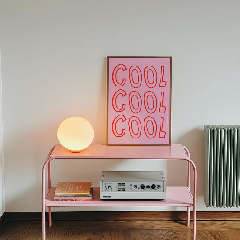May include: A pink metal side table with a round, glowing lamp and a framed print that says "COOL COOL COOL" in red. A stereo and books are on the table's lower shelf. A mint green radiator is visible in the background.