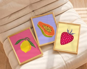 May include: Three rectangular wall art prints in thin gold frames. The left print features a yellow lemon on a pink background. The center print shows a sliced orange papaya on a light purple background. The right print displays a red strawberry on a pale yellow background.