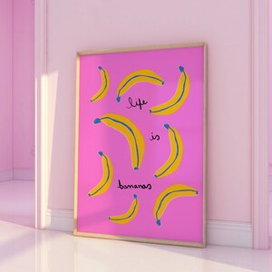 Może przedstawiać: R&oacute;żowy plakat w ramie z tekstem "life is bananas" w czarnych i niebieskich literach. Plakat przedstawia wz&oacute;r ż&oacute;łtych banan&oacute;w na r&oacute;żowym tle.