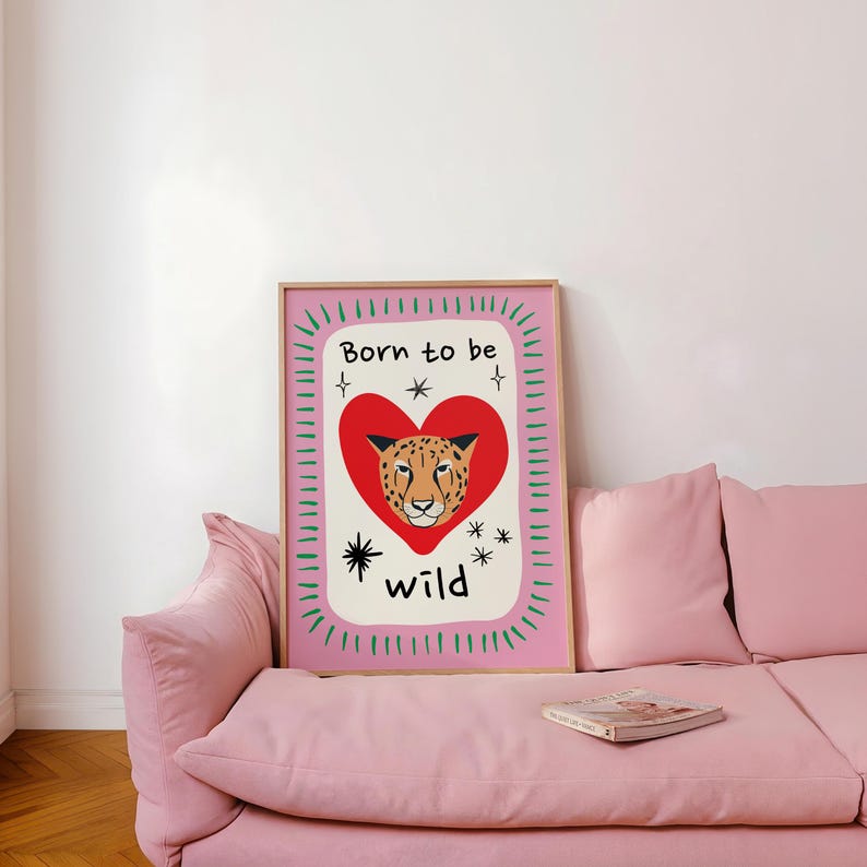 May include: A rectangular wall art print featuring a central red heart containing a cheetah face illustration. The text Born to be wild is printed in black above and below the heart. The design includes black star accents and a green dashed border on a pink background.