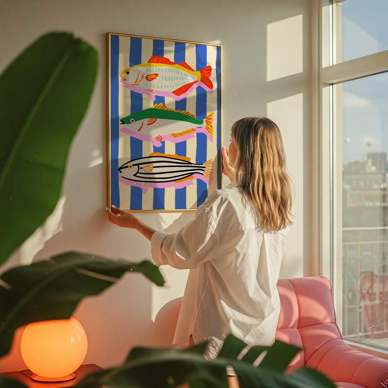 May include: A framed art print featuring three colorful fish illustrations against a blue and cream striped background. The fish have red, green, and pink accents. The artwork is in a gold frame.