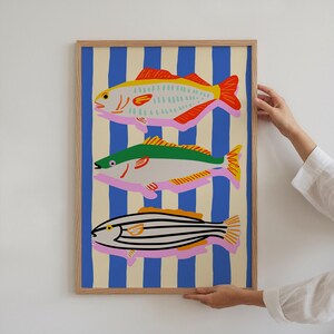 May include: A framed art print featuring three stylized fish illustrations. The fish are depicted in bright colors against a background of blue and cream vertical stripes. The fish have red, yellow, green, and black details.