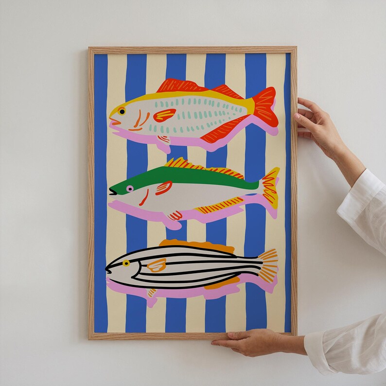 May include: A framed art print featuring three stylized fish illustrations. The fish are depicted in bright colors against a background of blue and cream vertical stripes. The fish have red, yellow, green, and black details.