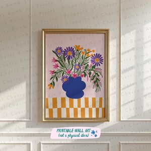以下が含まれることがあります： 青い花瓶に活けられた鮮やかな花の絵が額装されています。花は紫、オレンジ、ピンクで、薄いピンク色の背景に配置されています。花瓶は、黄色と白の市松模様のテーブルの上に置かれています。下部のテキストは「PRINTABLE WALL ART（not a physical item）」です。