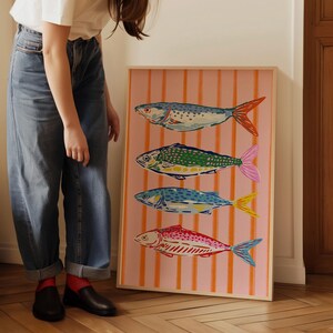 May include: A framed art print displaying four colourful fish illustrations. The fish are blue, green, and red, with orange and pink details. The background is pink with vertical orange stripes. The artwork is in a wooden frame.