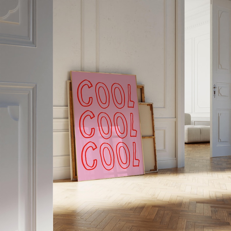 May include: A pink art print featuring the word "COOL" in red, repeated three times. The print is propped against a white wall in a room with a wooden floor. The room has an open doorway.