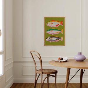 Puede incluir: Una impresi&oacute;n art&iacute;stica enmarcada con tres ilustraciones de peces estilizados. Los peces son de color rosa, amarillo, verde y negro sobre un fondo verde. La obra de arte se exhibe en una pared blanca en una habitaci&oacute;n con una mesa y una silla de madera.