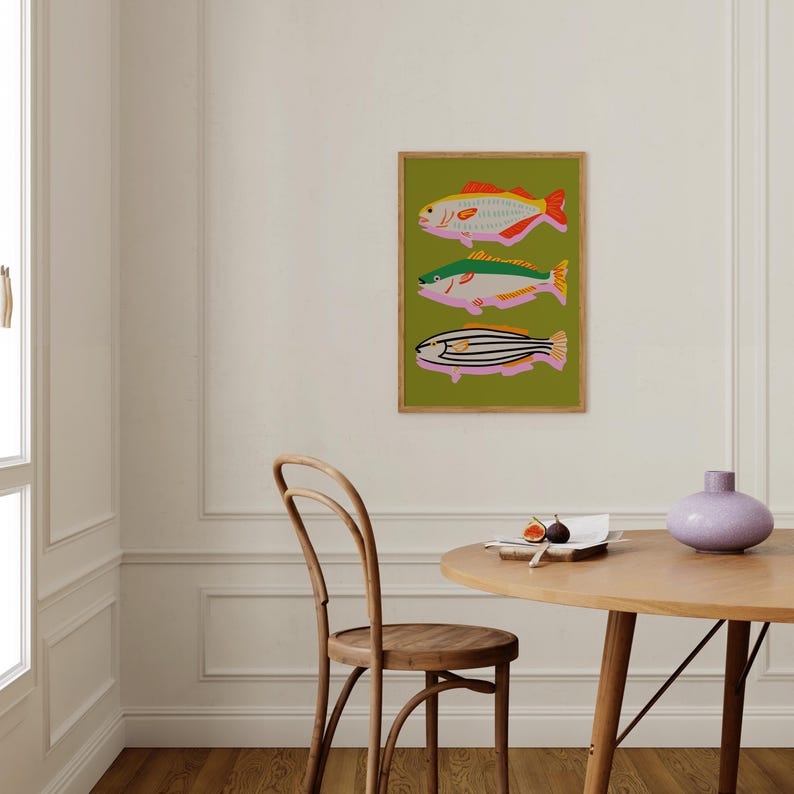 Puede incluir: Una impresi&oacute;n art&iacute;stica enmarcada con tres ilustraciones de peces estilizados. Los peces son de color rosa, amarillo, verde y negro sobre un fondo verde. La obra de arte se exhibe en una pared blanca en una habitaci&oacute;n con una mesa y una silla de madera.