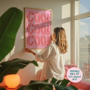 May include: A framed pink wall art print with the word "COOL" repeated three times in red. The print is being held up by a person. The print is in a gold frame. The text "PRINTABLE WALL ART (not a physical item)" is in the bottom right corner.