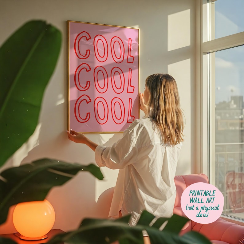 May include: A framed pink wall art print with the word "COOL" repeated three times in red. The print is being held up by a person. The print is in a gold frame. The text "PRINTABLE WALL ART (not a physical item)" is in the bottom right corner.