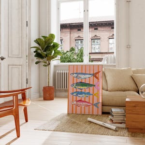 May include: A framed art print depicting four colourful fish designs on a pink striped background. The artwork is displayed in a well-lit room with a large window, a potted plant, and a wooden chair. The fish are in blue, green, yellow, and red.
