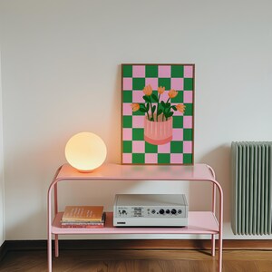 May include: A pink metal side table with a round, glowing lamp and a framed artwork. The artwork features a pink and green checkered background with a vase of orange flowers. A stereo and books are on the lower shelf.