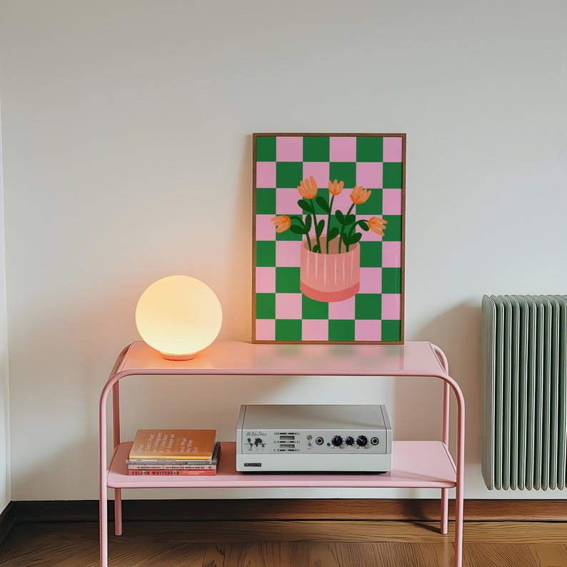 May include: A pink metal side table with a round, glowing lamp and a framed artwork. The artwork features a pink and green checkered background with a vase of orange flowers. A stereo and books are on the lower shelf.