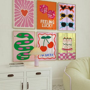 May include: Six framed art prints with vibrant, playful designs. The prints showcase a heart-shaped flower, dice with the text "Feeling Lucky", sunglasses, a green snake, cherries with the text "Ma Ch&eacute;rie", and a cake. Gold frames.