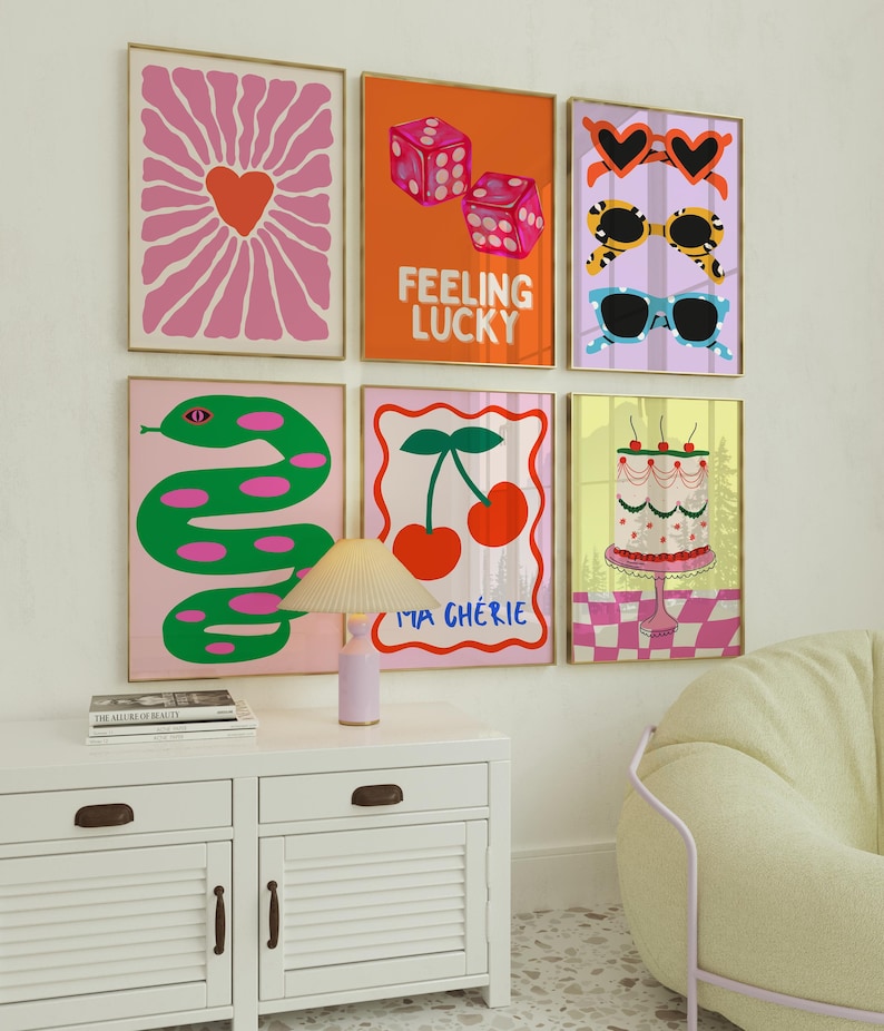 May include: Six framed art prints with vibrant, playful designs. The prints showcase a heart-shaped flower, dice with the text "Feeling Lucky", sunglasses, a green snake, cherries with the text "Ma Ch&eacute;rie", and a cake. Gold frames.