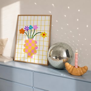 May include: A framed wall art print featuring a yellow grid background with a pink and yellow checkered vase holding colorful flowers, a silver disco ball, and a light pink spiral candle in a tan croissant-shaped candle holder.