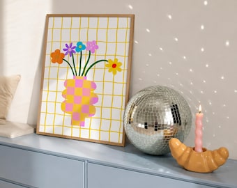 May include: A framed wall art print featuring a yellow grid background with a pink and yellow checkered vase holding colorful flowers, a silver disco ball, and a light pink spiral candle in a tan croissant-shaped candle holder.