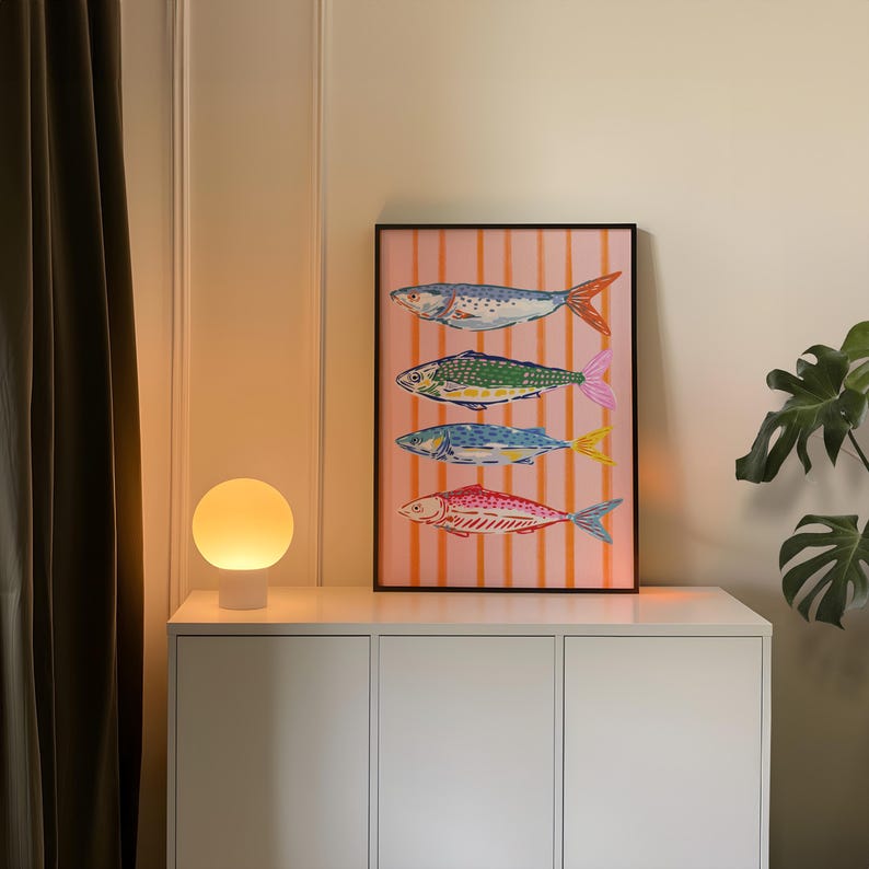May include: A framed art print displaying four illustrated fish in various colours, set against a pink and white striped backdrop. The fish feature detailed scales and fins. The artwork is displayed on a white cabinet, with a round lamp to the left.