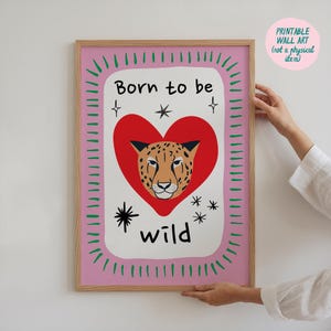 May include: A framed wall art print featuring a central red heart containing a leopard face illustration. The text Born to be wild is printed in black. The design includes black star accents and a pink border with green dash marks.