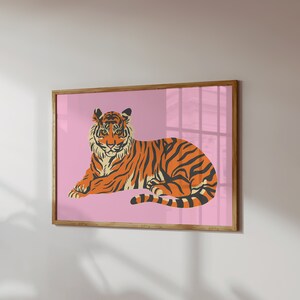 May include: A framed print of a tiger lying down on a pink background. The tiger is orange and black with white stripes.