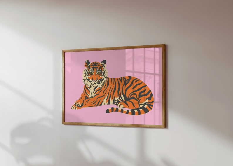 May include: A framed print of a tiger lying down on a pink background. The tiger is orange and black with white stripes.