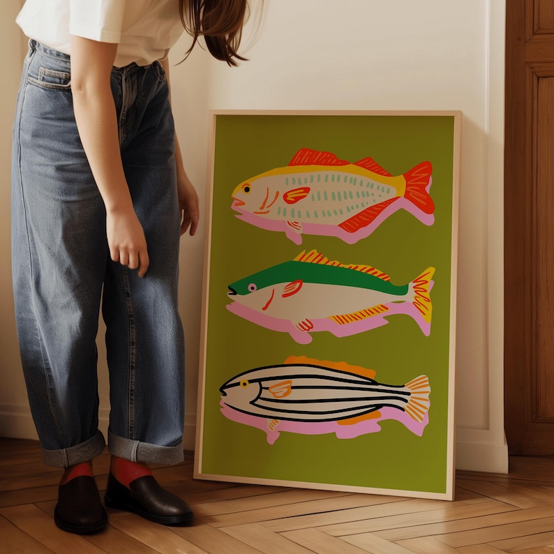 Puede incluir: Un cuadro enmarcado con tres ilustraciones de peces estilizados. Los peces son de color rojo, amarillo, verde y negro, sobre un fondo verde claro. La obra de arte se presenta en un marco de madera.