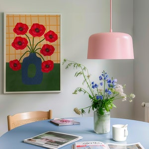 May include: A blue vase with red poppy flowers in a white frame on a white wall. A pink pendant light hangs above a blue table with a bouquet of flowers in a clear glass vase. There are magazines on the table.