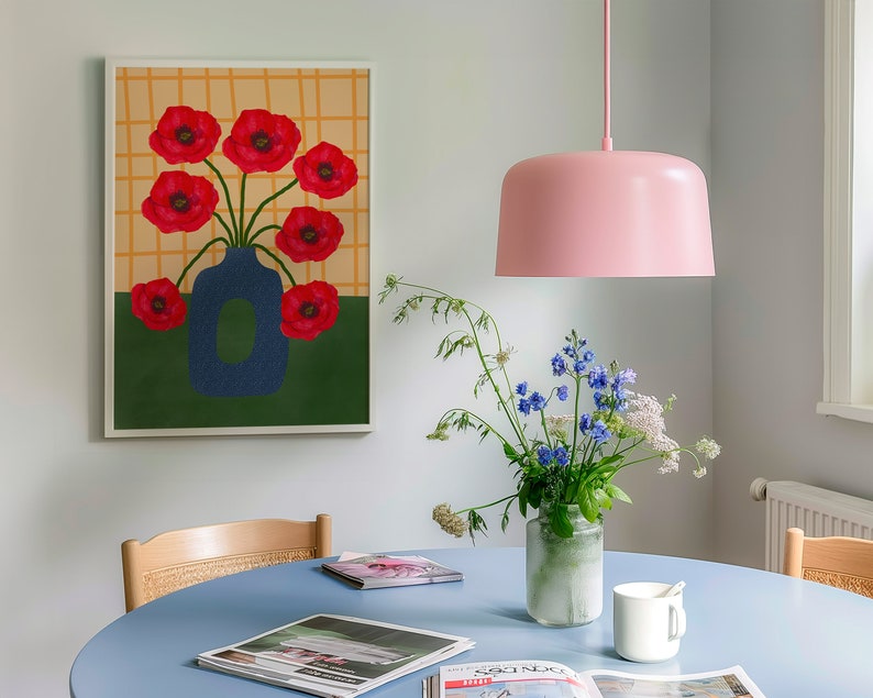 May include: A blue vase with red poppy flowers in a white frame on a white wall. A pink pendant light hangs above a blue table with a bouquet of flowers in a clear glass vase. There are magazines on the table.