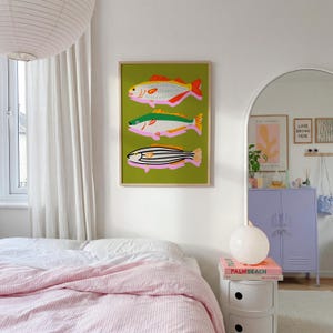 Puede incluir: Una impresi&oacute;n art&iacute;stica enmarcada con tres ilustraciones estilizadas de peces sobre un fondo verde. Los peces son en tonos rosa, amarillo, naranja y negro, con un marco dorado. La obra de arte se exhibe en una pared blanca en un dormitorio.