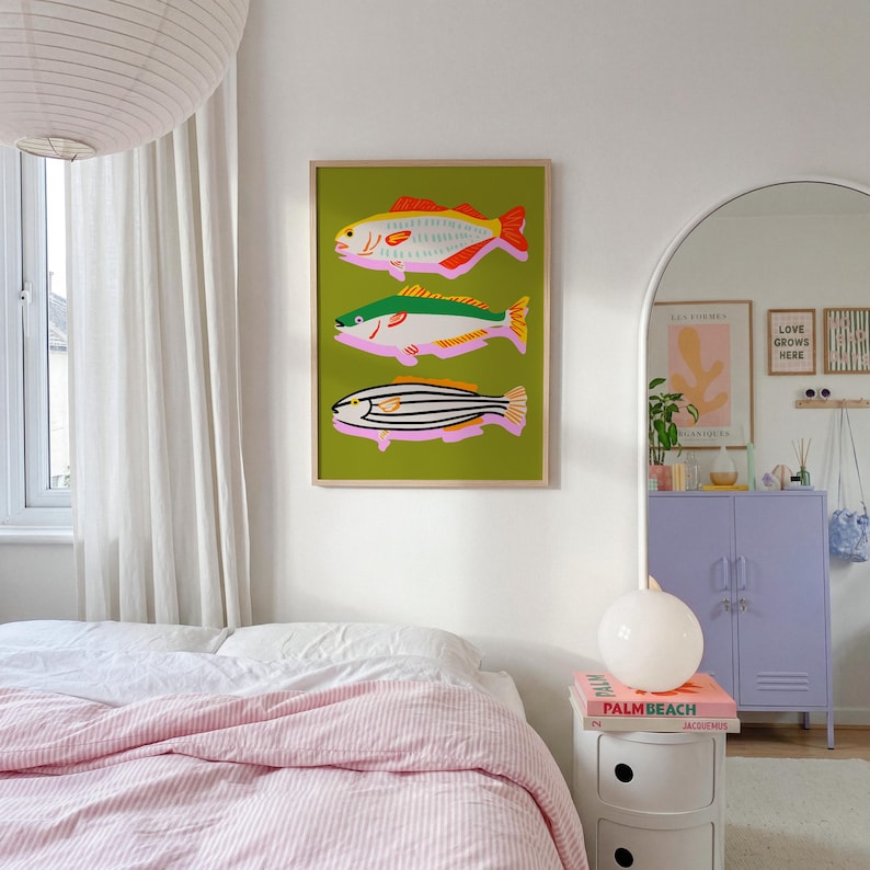 Puede incluir: Una impresi&oacute;n art&iacute;stica enmarcada con tres ilustraciones estilizadas de peces sobre un fondo verde. Los peces son en tonos rosa, amarillo, naranja y negro, con un marco dorado. La obra de arte se exhibe en una pared blanca en un dormitorio.