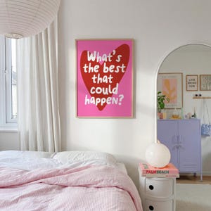 May include: A framed pink art print with a red heart and the text "What's the best that could happen?" is displayed on a white wall. The room features a bed with pink striped bedding, a white nightstand, and a purple cabinet.