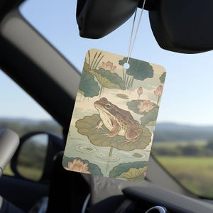 May include: Rectangular air freshener featuring a frog on a lily pad in a pond with lotus flowers. The air freshener has a light green and beige color scheme, with a string for hanging. The background shows a blue sky and a car interior.