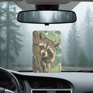 May include: Rectangular car air freshener featuring a raccoon peeking from behind a tree. The design has a Japanese art style with a green and brown color palette. The air freshener hangs from a string, with a forest background visible through the car's windshield.