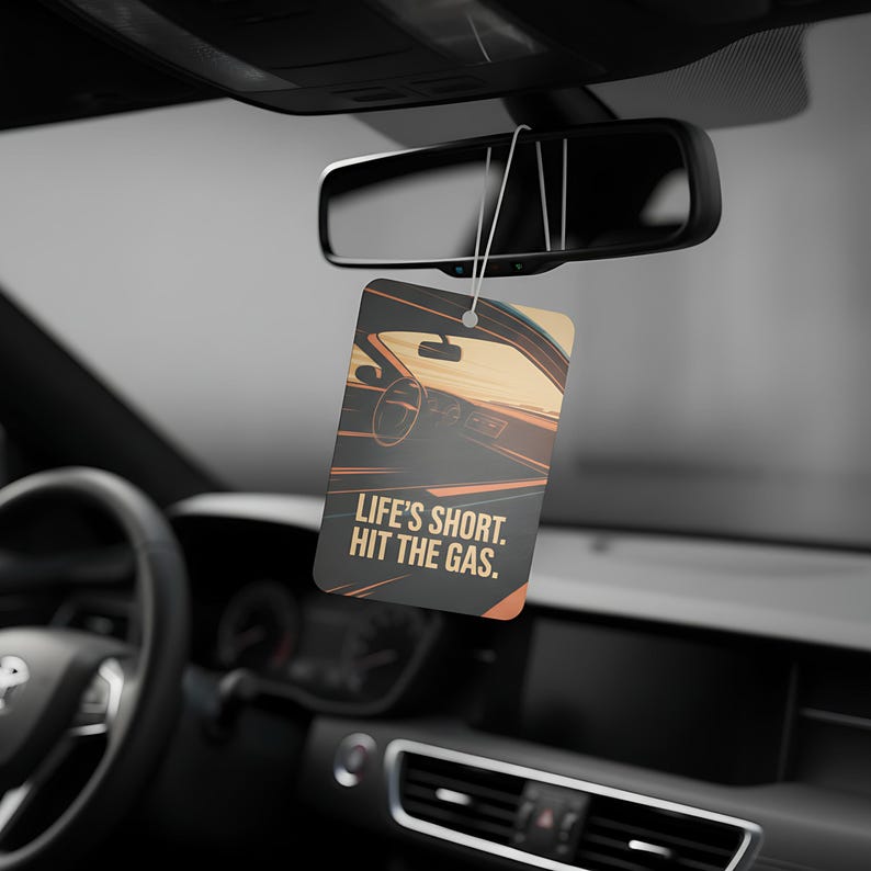May include: A rectangular car air freshener with a car interior graphic and the text "LIFE'S SHORT. HIT THE GAS." is hanging from a car's rearview mirror. The air freshener has a black background with orange and tan accents.
