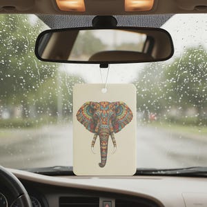 May include: Rectangular car air freshener with a colorful elephant design. The elephant features intricate patterns in shades of red, blue, green, and yellow. The air freshener hangs from a string in front of a car's rearview mirror.