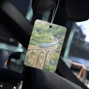 May include: Rectangular air freshener with a watercolor painting of a blue train crossing a stone bridge. The bridge arches over a lush green landscape. The air freshener hangs from a white string.