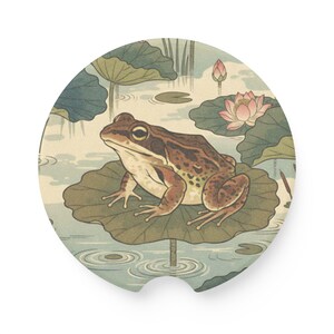 May include: Round car coaster featuring a detailed illustration of a frog sitting on a lily pad in a pond. The frog is brown and tan, with a light green lily pad. The background includes water, lily pads, and a pink lotus flower.