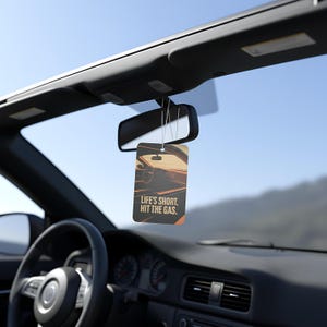 May include: Rectangular car air freshener with a car interior graphic and the text "LIFE'S SHORT. HIT THE GAS." The air freshener hangs from the rearview mirror in a car. The car's dashboard and steering wheel are visible.