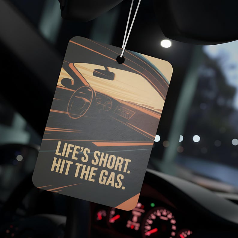 May include: Rectangular car air freshener with a graphic of a car interior in shades of brown, orange, and black. The text "LIFE'S SHORT. HIT THE GAS." is printed in white. The air freshener hangs from a white string.