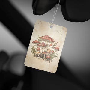 May include: A rectangular tag with a watercolor illustration of mushrooms. The mushrooms have red caps with white spots and are surrounded by green foliage. The tag has a beige background and a small hole at the top for hanging.