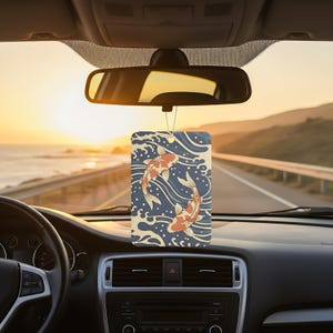 May include: Rectangular car air freshener with a Japanese koi fish design. The air freshener features two koi fish in shades of orange and white, swimming in a blue and cream-colored wave pattern. It hangs from a string attached to the rearview mirror.
