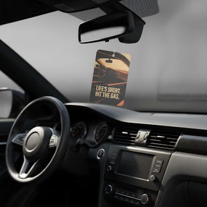 May include: Rectangular car air freshener with a car interior graphic and the text "LIFE'S SHORT. HIT THE GAS." The air freshener hangs from a rearview mirror in a car. The car's interior is black.