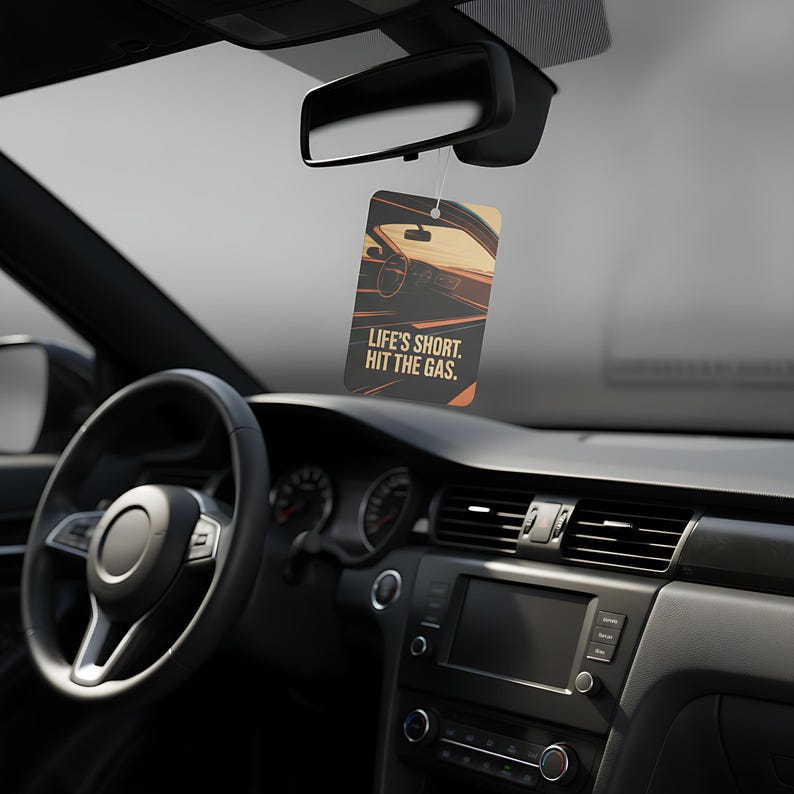 May include: Rectangular car air freshener with a car interior graphic and the text "LIFE'S SHORT. HIT THE GAS." The air freshener hangs from a rearview mirror in a car. The car's interior is black.