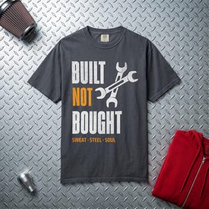May include: A charcoal gray short-sleeve t-shirt featuring the white and orange text BUILT NOT BOUGHT with a graphic of two crossed wrenches. Below the main text, the words SWEAT STEEL SOUL are printed in orange. The shirt has a crew neckline.