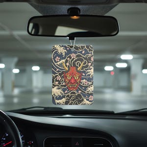 May include: Rectangular car air freshener with a Japanese demon mask design. The mask is red with gold horns, set against a navy blue background with white waves, pink cherry blossoms, and black clouds. The air freshener hangs from a car's rearview mirror.