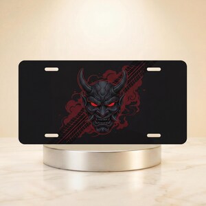 May include: Black license plate featuring a detailed illustration of a Japanese Oni mask with red eyes and horns. Red tire tracks and smoke-like designs are on the right side of the plate.