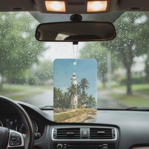 May include: Rectangular car air freshener featuring a coastal scene with a white lighthouse, palm trees, and a blue sky. The air freshener hangs from a car's rearview mirror, with a rainy outdoor backdrop.