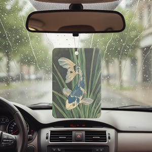 May include: Rectangular car air freshener featuring a koi fish design. The artwork depicts a blue, white, and gold koi fish swimming among green reeds against a dark background. The air freshener hangs from a string, visible through a car windshield.