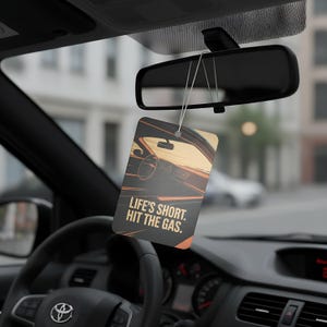 May include: Rectangular car air freshener with a car interior graphic and the text "LIFE'S SHORT. HIT THE GAS." The air freshener hangs from the rearview mirror in a car. The graphic features shades of orange, brown, and black.