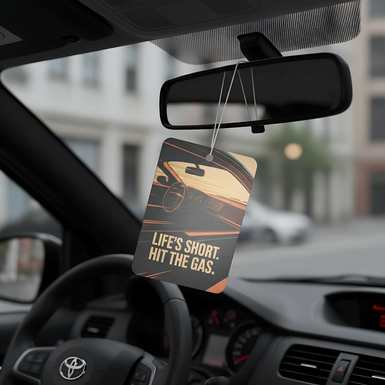 May include: Rectangular car air freshener with a car interior graphic and the text "LIFE'S SHORT. HIT THE GAS." The air freshener hangs from the rearview mirror in a car. The graphic features shades of orange, brown, and black.